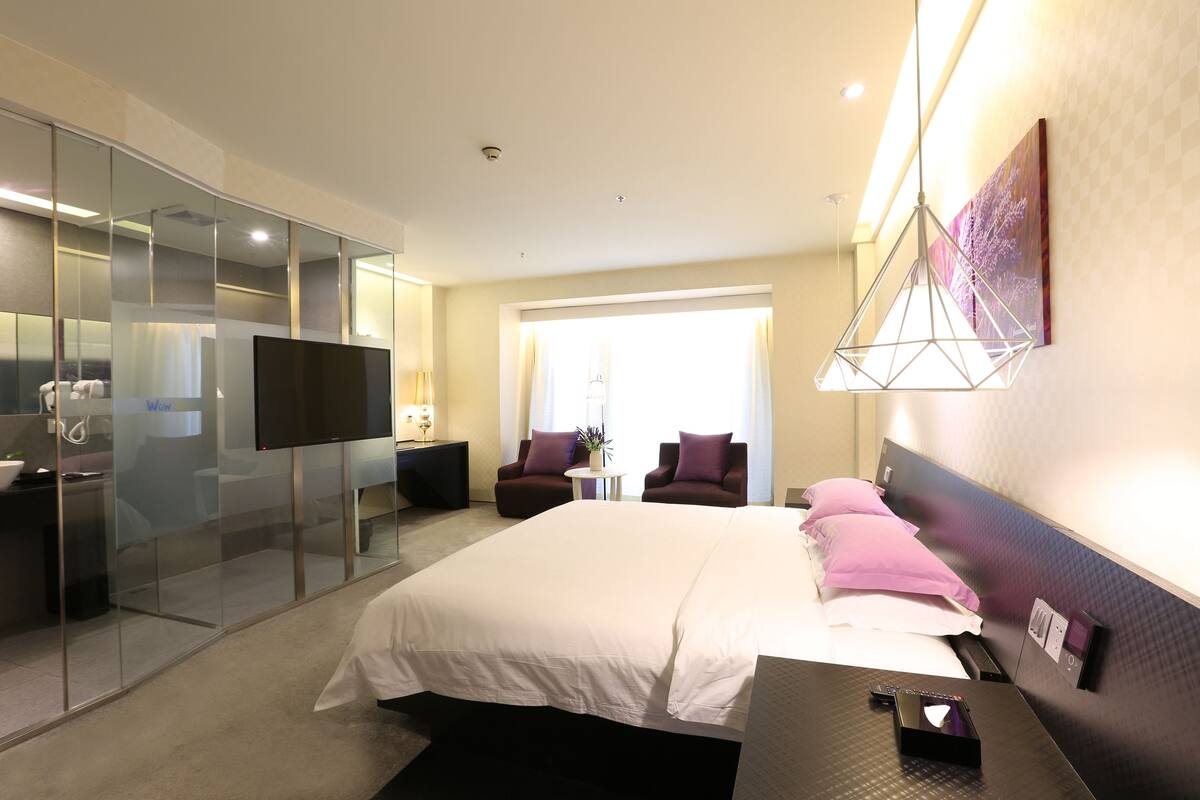 Una habitación de hotel moderna con una cama grande, televisión, escritorio y un panel de vidrio que separa la habitación del baño.