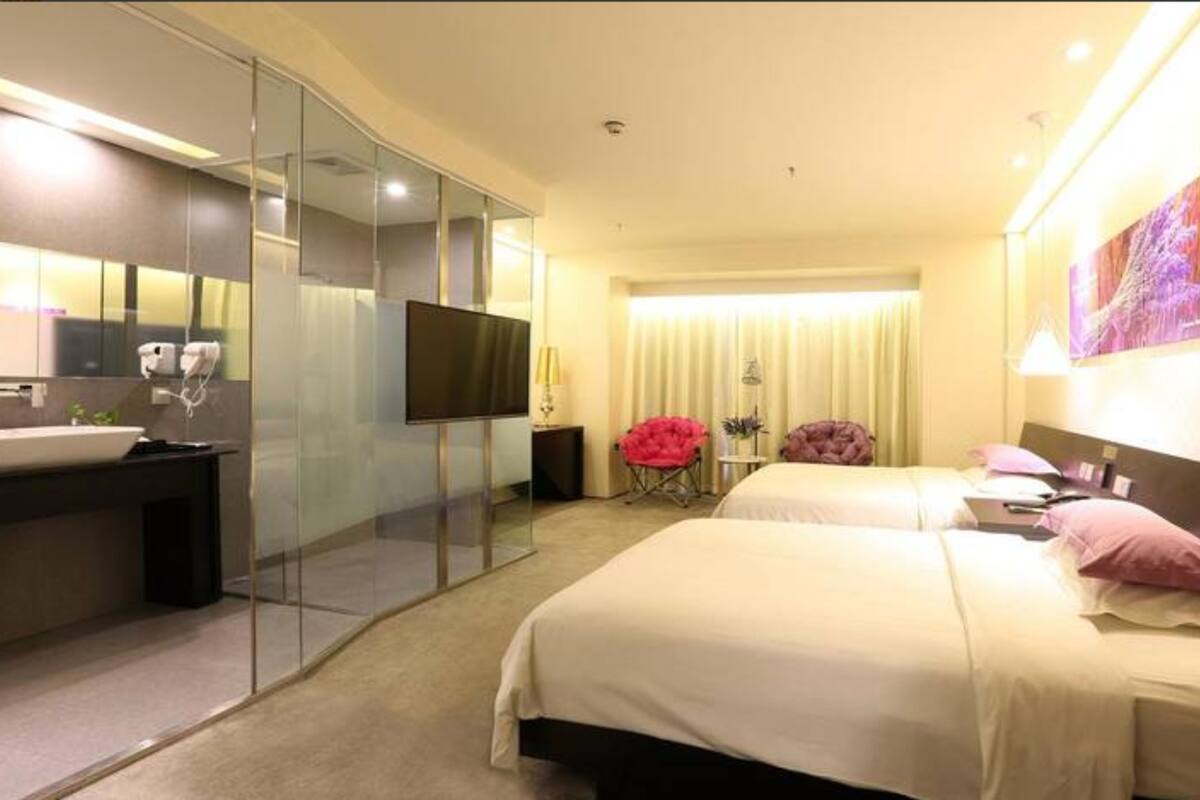 Habitación de hotel con baño de vidrio, televisor de pantalla plana, cama con dos almohadas y una pequeña mesa con lámpara.