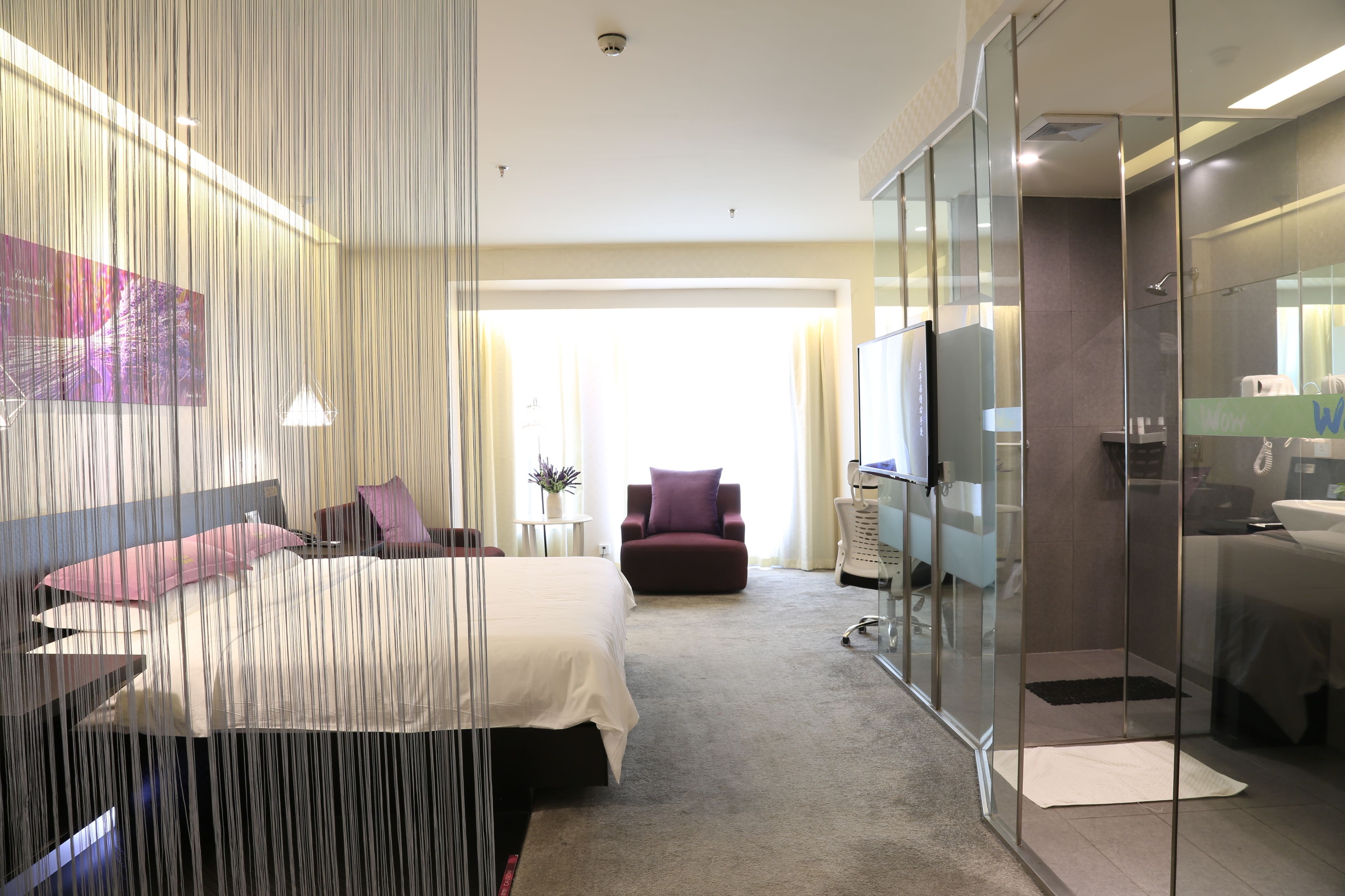 A modern hotel room with a large bed, a seating area with a chair, a glass-enclosed shower, and a television.
