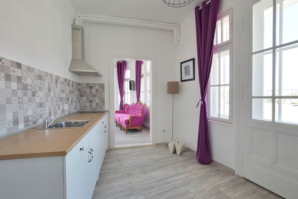 A modern kitchen with a wooden countertop, a built-in sink, and a patterned backsplash. A pink sofa is visible in the background, leading to a living area. - Hostel Musala (Varna)