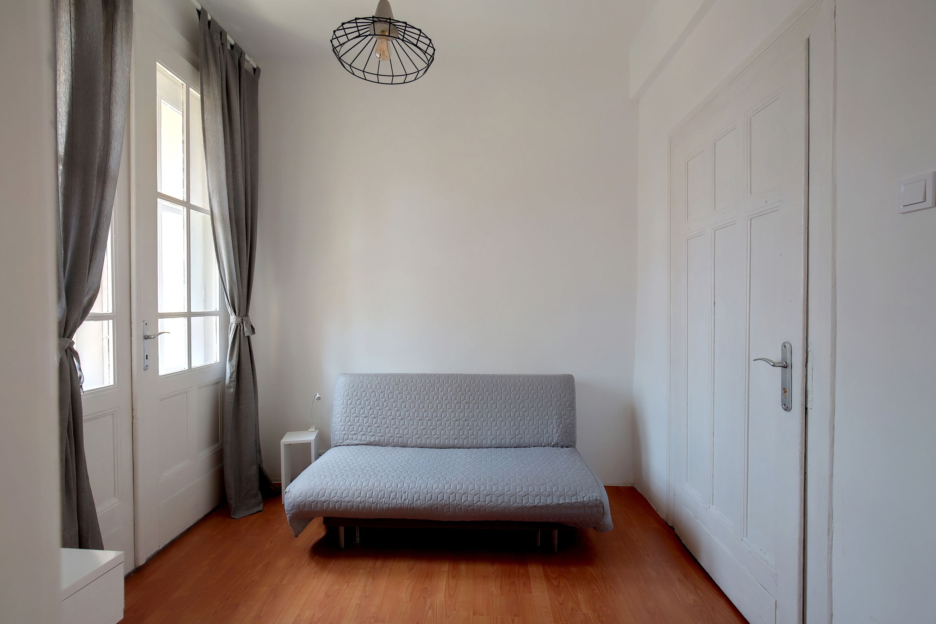 A room with a sofa bed, a window with curtains, a door, and a hanging light fixture.