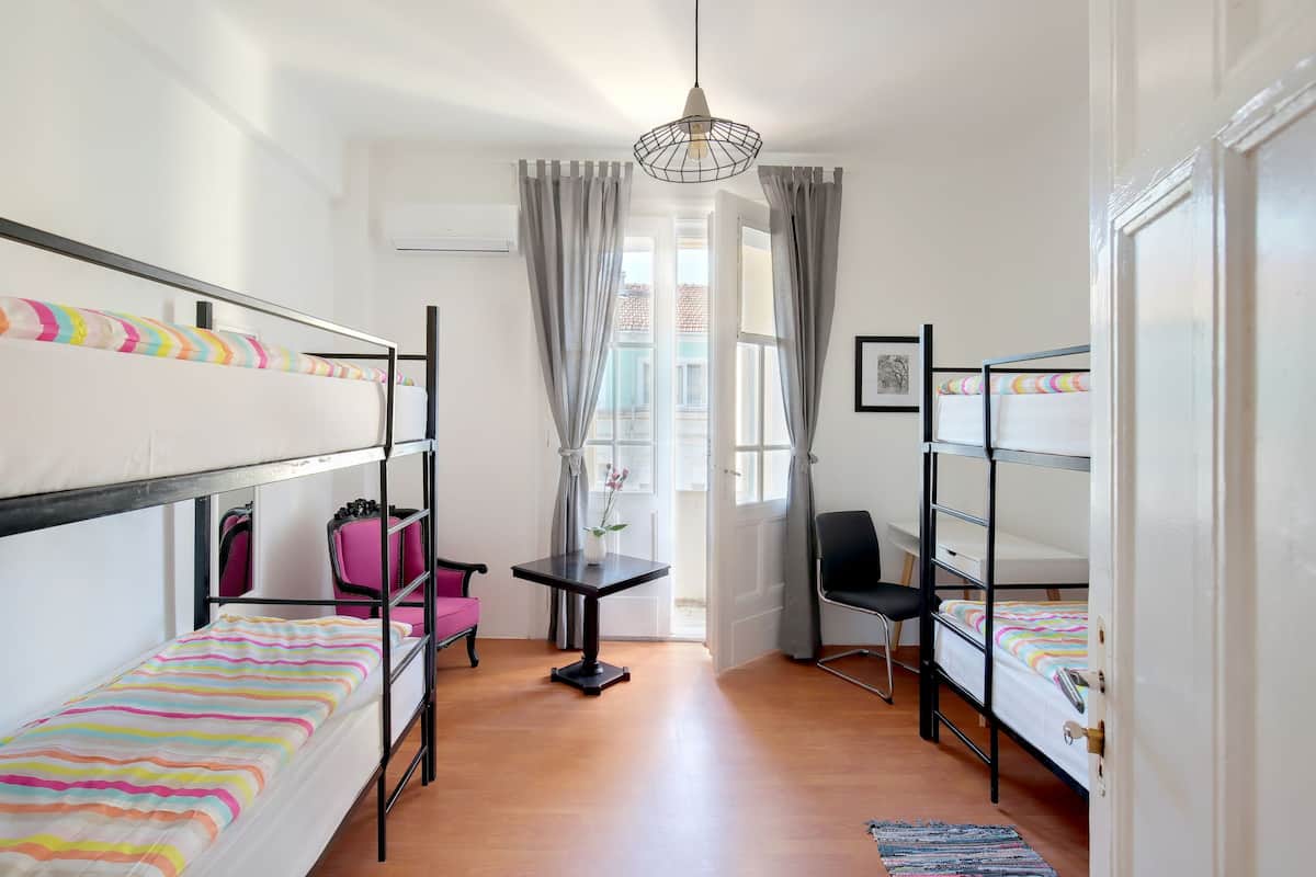 A dormitory room with bunk beds, a small table, a chair, and a view of the outside.
