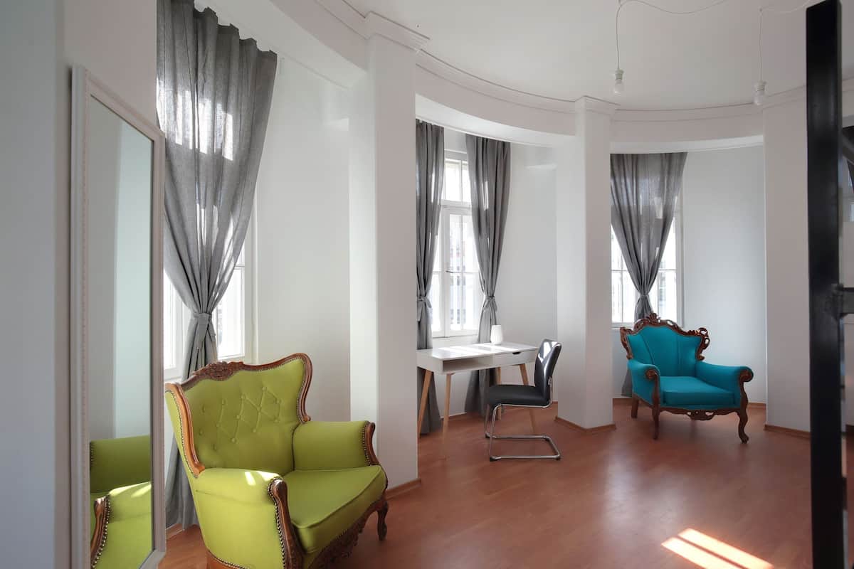 A room with a wooden floor, a green armchair, a blue armchair, a white desk, and a mirror.