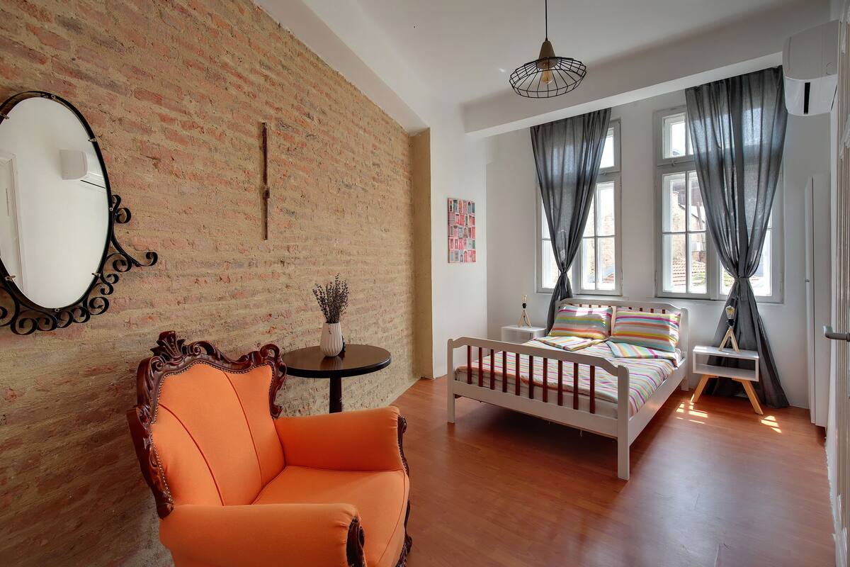 A room with a brick wall, a white bed with colorful bedding, a wooden chair, a round table with a vase, and a hanging light fixture.