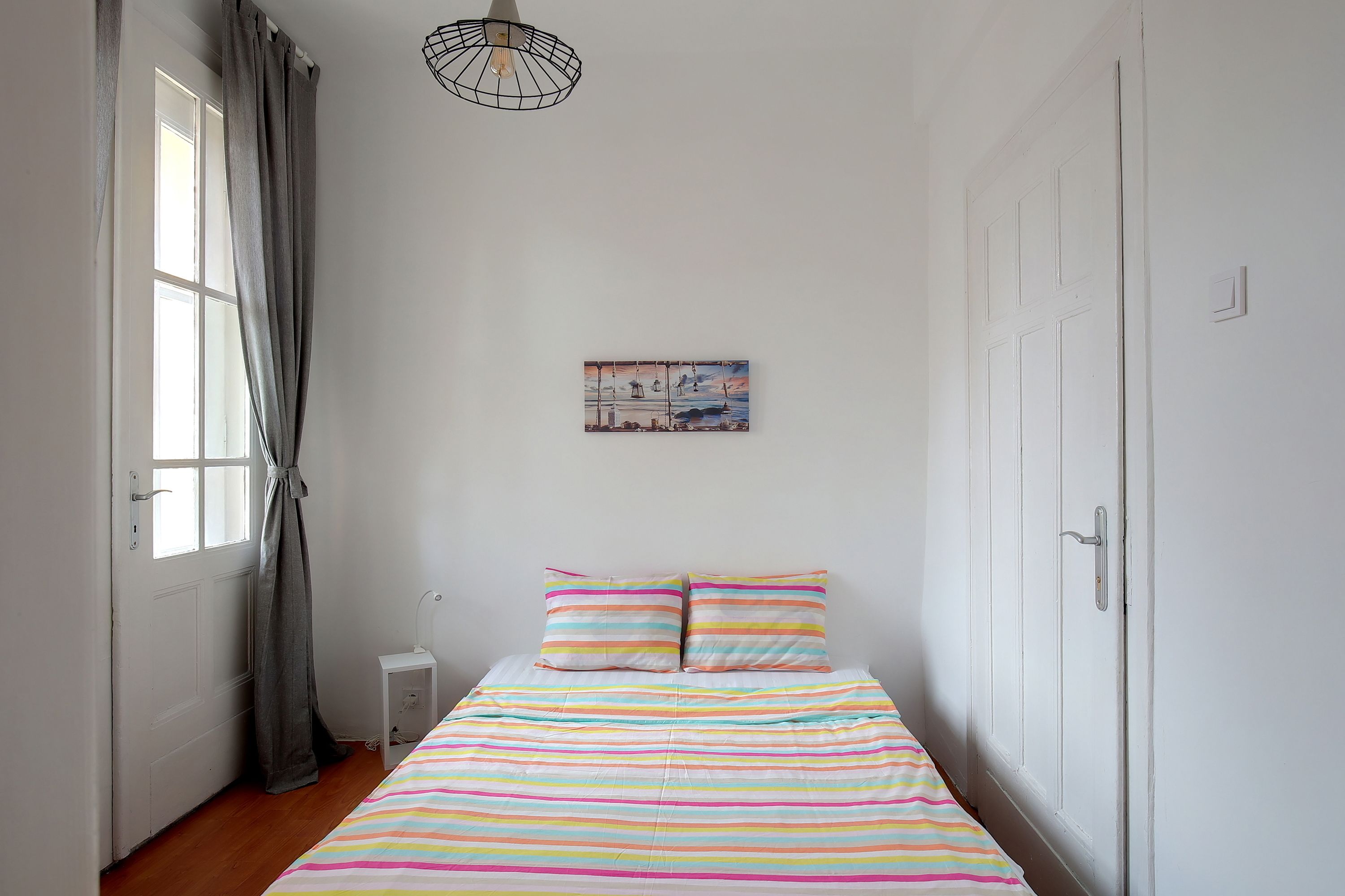A bedroom with a bed, a window with curtains, a wall-mounted light fixture, and a door.