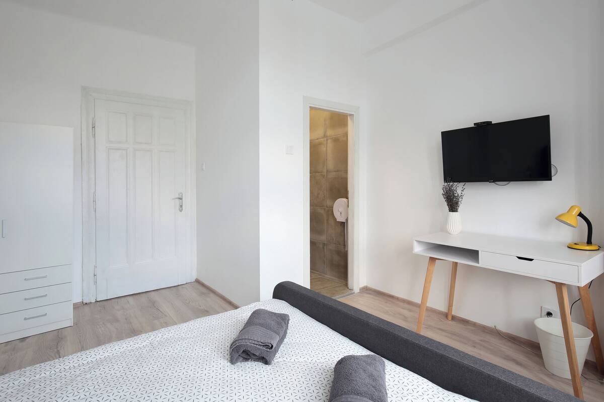 A modern bedroom with a bed, a desk, a TV, and a bathroom visible through an open door.