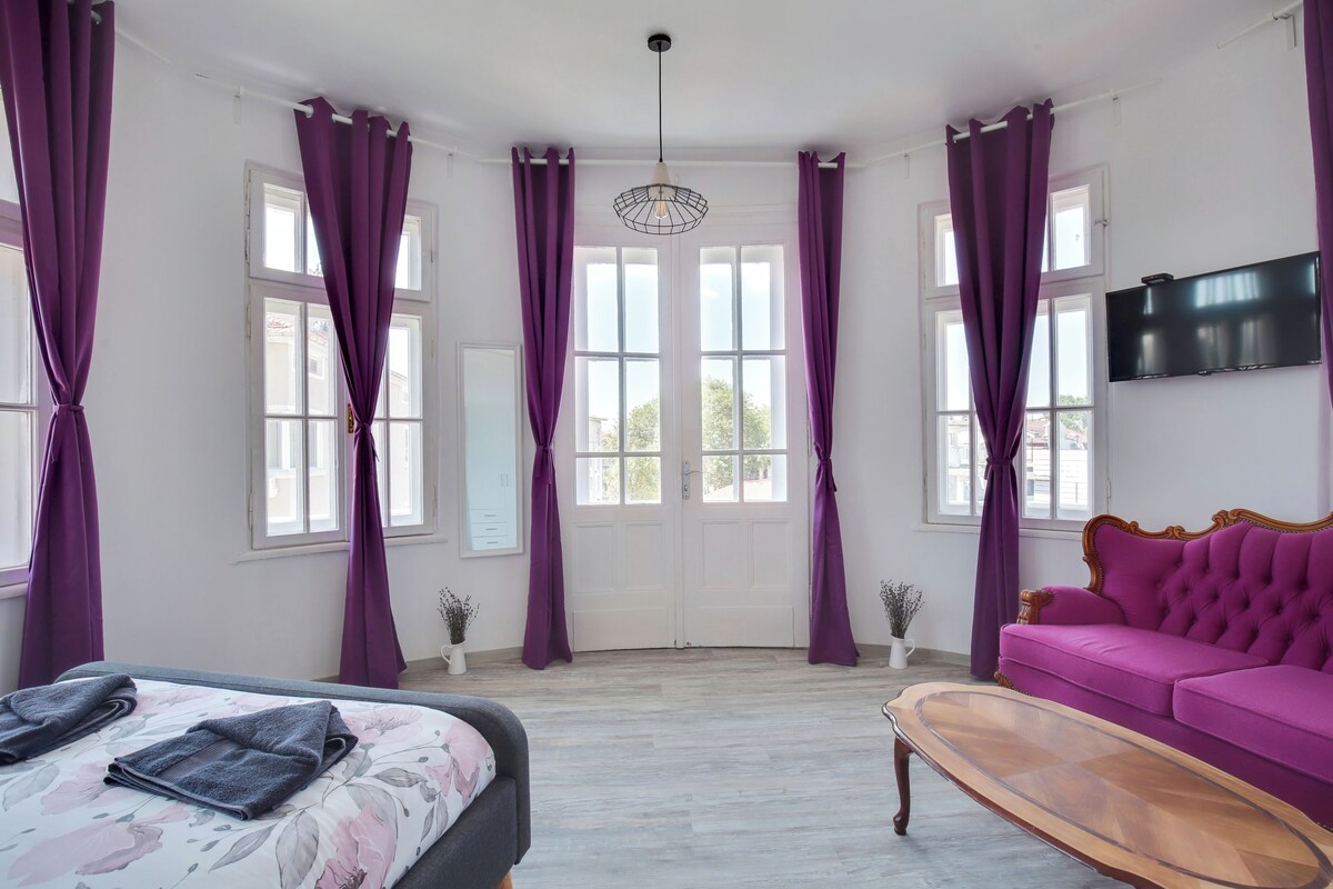 A room with a bed, a purple sofa, a TV, and large windows with purple curtains.