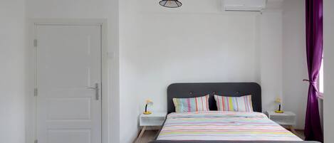 A bedroom with a bed, a nightstand, a lamp, a door, and an air conditioning unit.