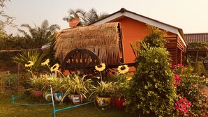 A rustic house with a thatched roof, surrounded by a garden with sunflowers and flowering plants. - Bussaracum Hotel (Tha Muang)
