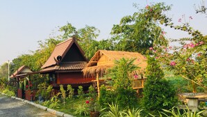A traditional wooden house with a thatched roof, surrounded by lush greenery and flowering plants.
