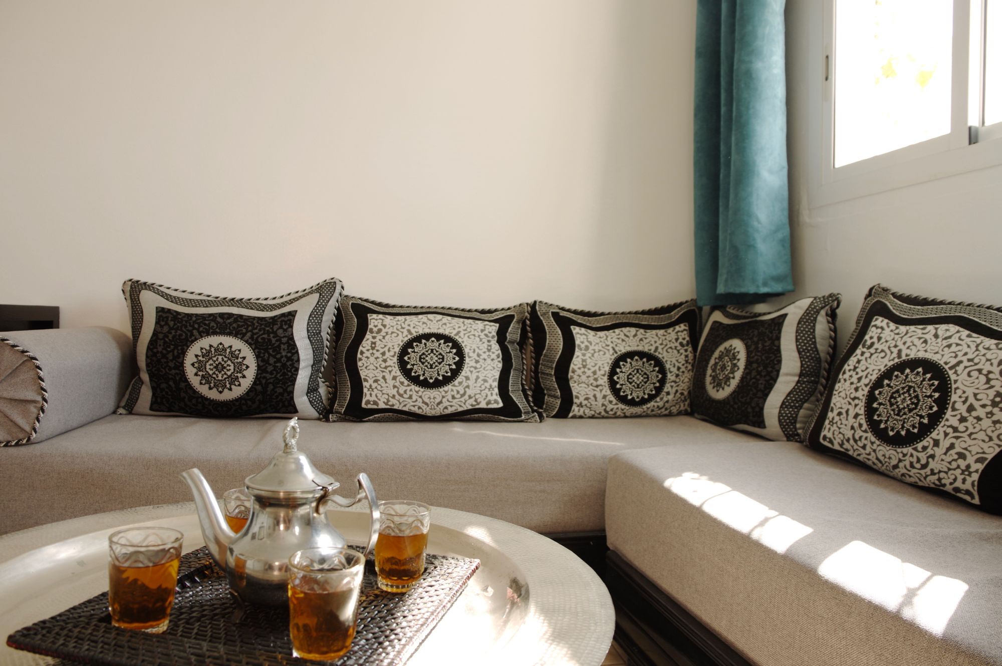 Un sofá con cojines estampados, una bandeja con tetera y vasos, y una ventana con cortina.