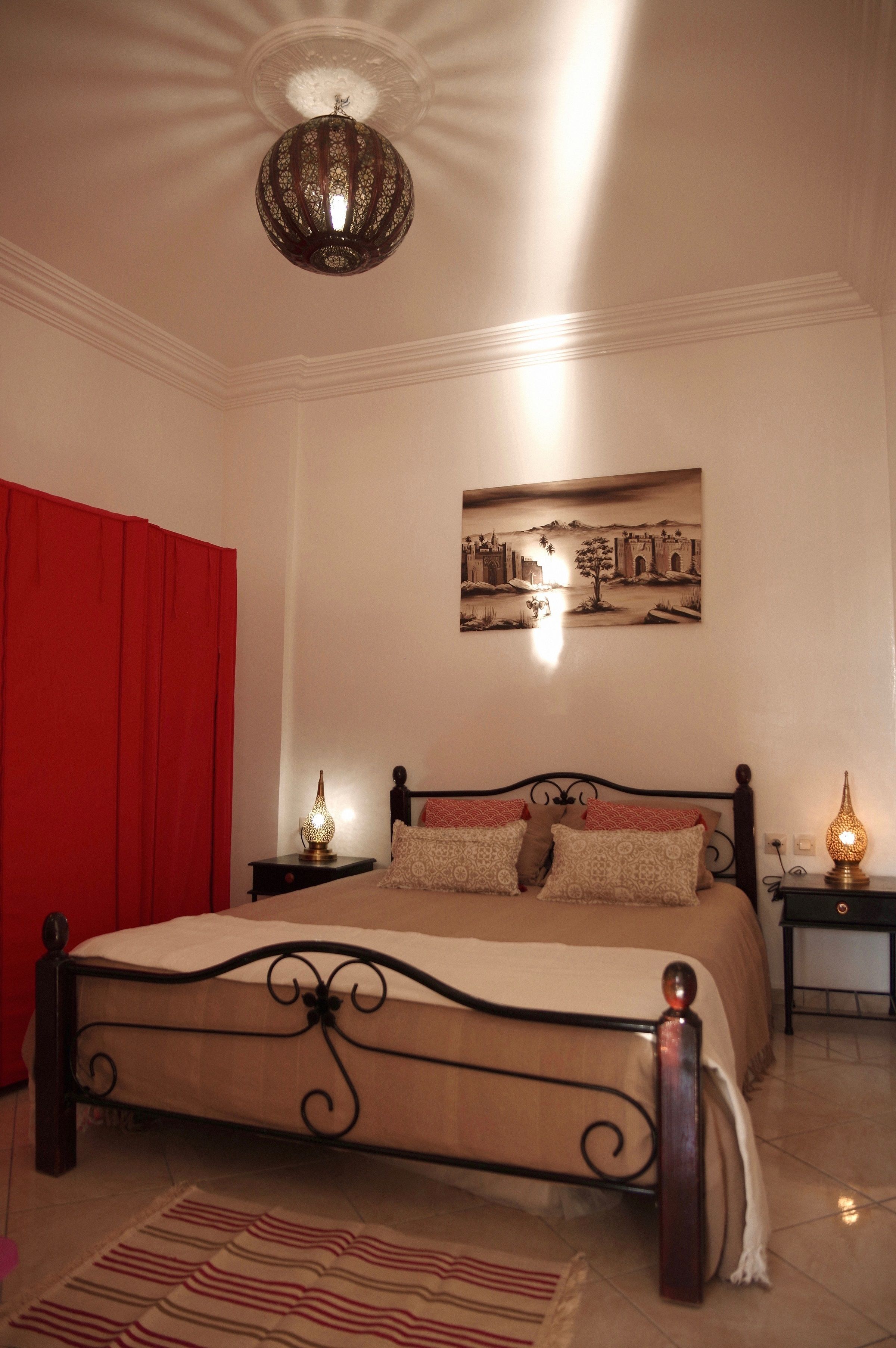 Une chambre à coucher avec un lit, un rideau rouge, une lampe fixée au mur, une table de chevet et un tableau.