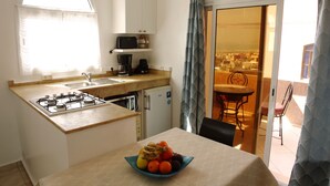 Eine kompakte Küche mit Gasherd, Kaffeemaschine und kleinem Kühlschrank. Vor der Essecke steht eine Schale mit Obst. Eine Glastür öffnet sich zu einem Balkon mit Blick auf die Stadt.