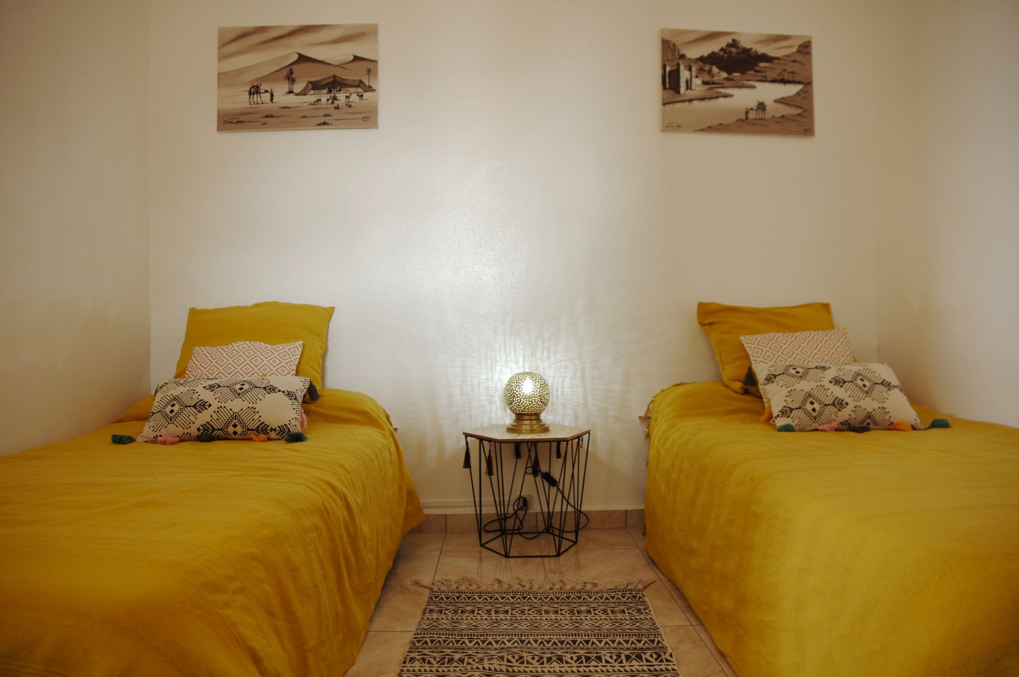 Una habitación con dos camas, cada una con ropa de cama amarilla y almohadas estampadas. Entre las camas hay una mesita redonda con una lámpara. Las paredes son lisas y hay dos cuadros enmarcados sobre las camas.