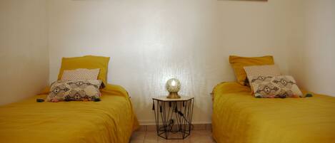 Una habitación con dos camas, cada una con ropa de cama amarilla y almohadas estampadas. Entre las camas hay una mesita redonda con una lámpara. Las paredes son lisas y hay dos cuadros enmarcados sobre las camas.
