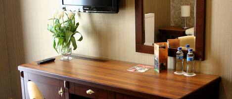 A hotel room with a wooden desk, a chair, a flat-screen TV, a mirror, a vase with flowers, and two water bottles.
