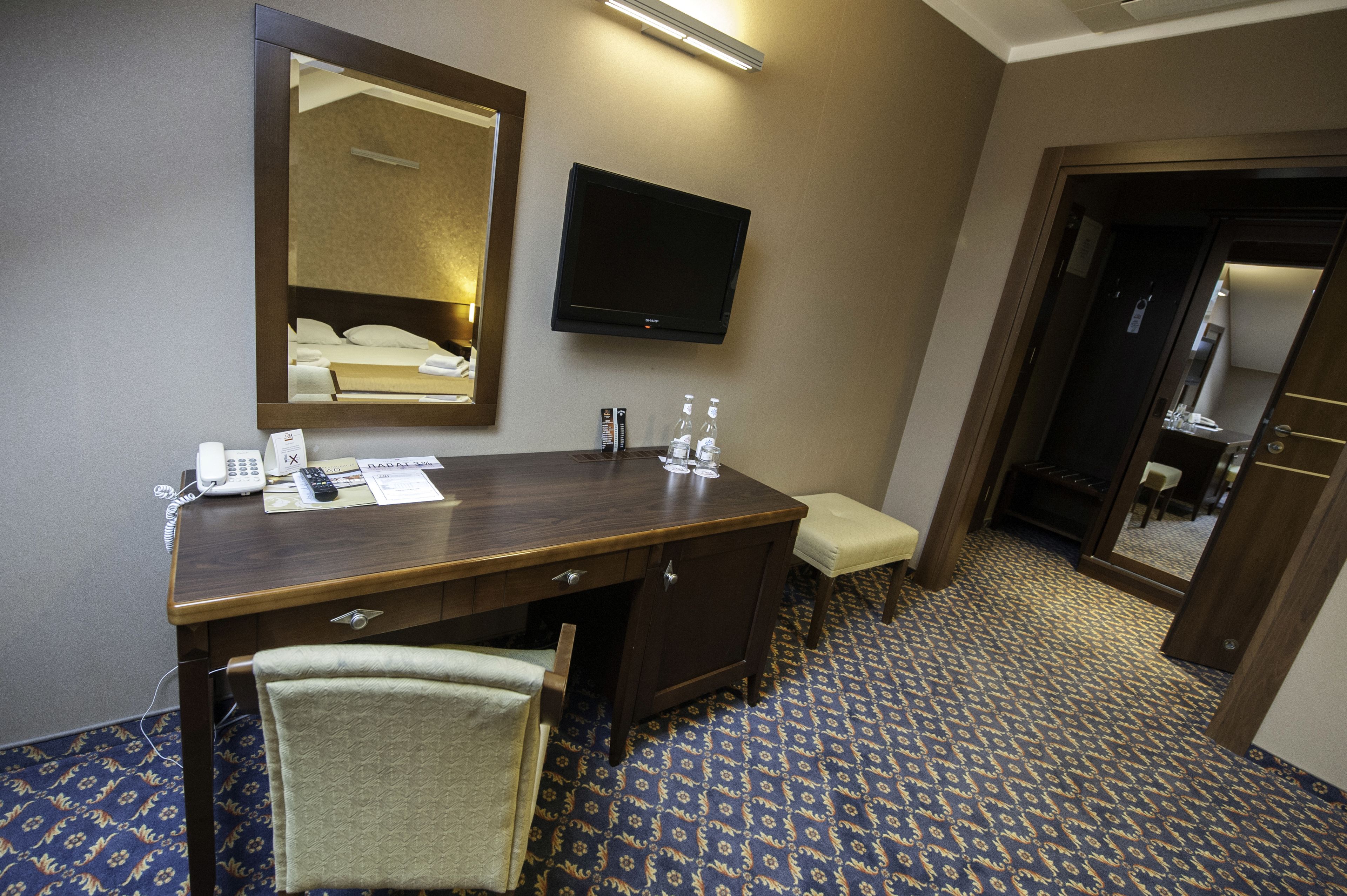Ein Hotelzimmer mit einem Holztisch, einem Spiegel, einem Flachbildfernseher, einem Telefon, einem Sessel und einem gemusterten Teppich.