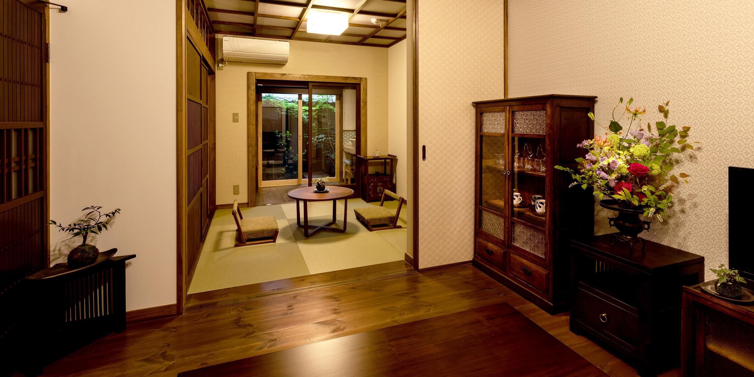 Une pièce de style japonais traditionnel, avec un sol recouvert de tatamis, une porte coulissante donnant sur un jardin, un meuble en bois aux portes vitrées et une petite table accompagnée d’une chaise.