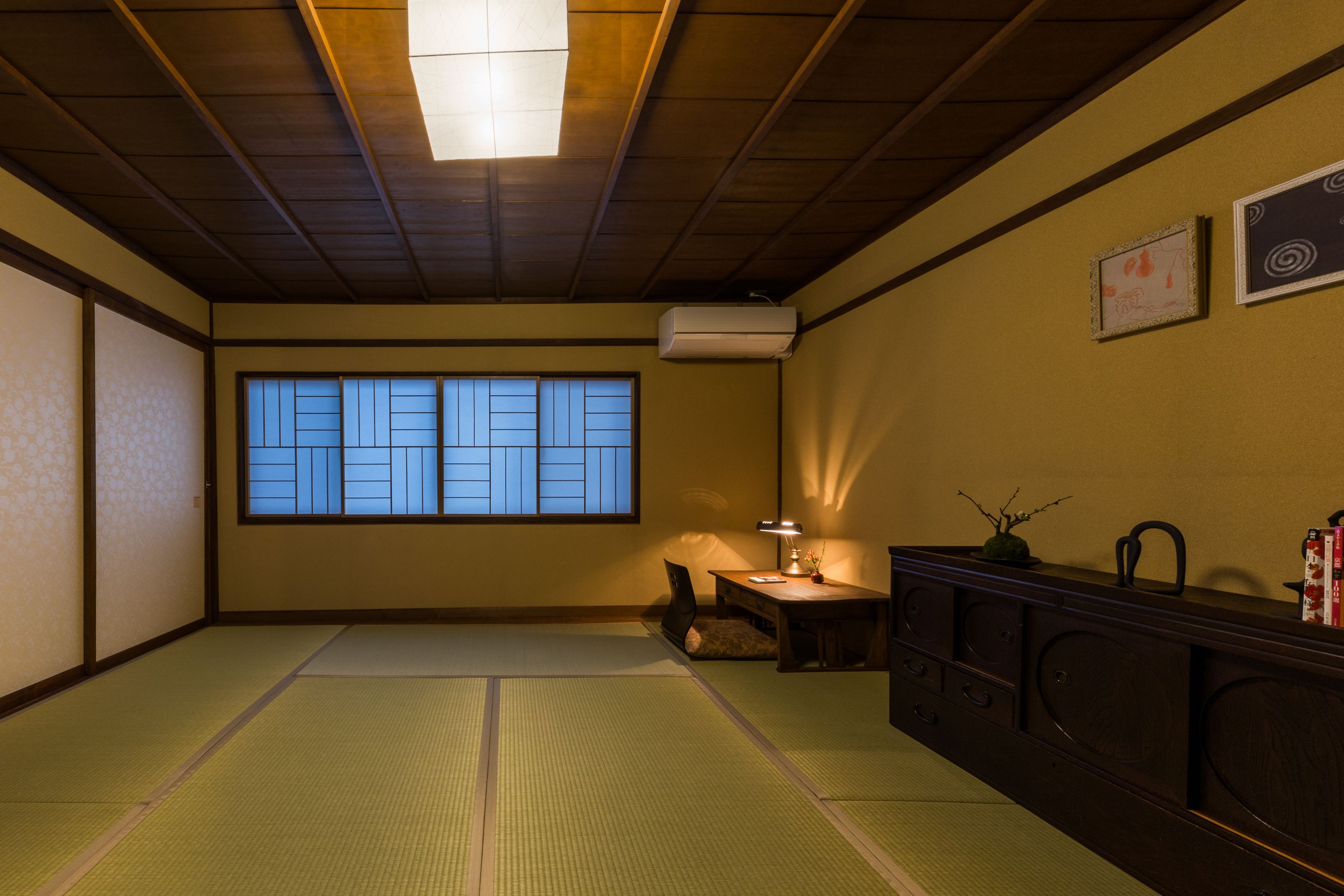Una habitación japonesa tradicional con suelo de tatami, una puerta corredera, un escritorio con lámpara y un aparador con objetos decorativos.