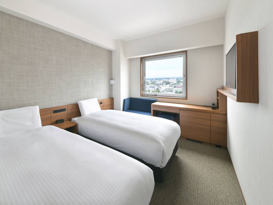Habitación de hotel con dos camas, un escritorio, una silla, un ventanal grande con vista a la ciudad y un televisor de pared.