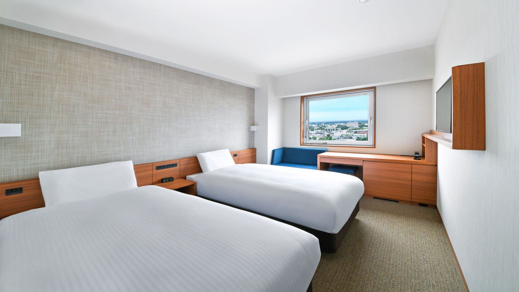 A hotel room with two beds, a desk, a chair, a large window with a city view, and a TV mounted on the wall.