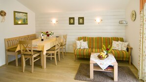 A dining area with a wooden table and chairs, a sofa with a striped cushion, and a small table with a fruit bowl and flowers. - Ferienwohnungen Christl (Reit im Winkl)