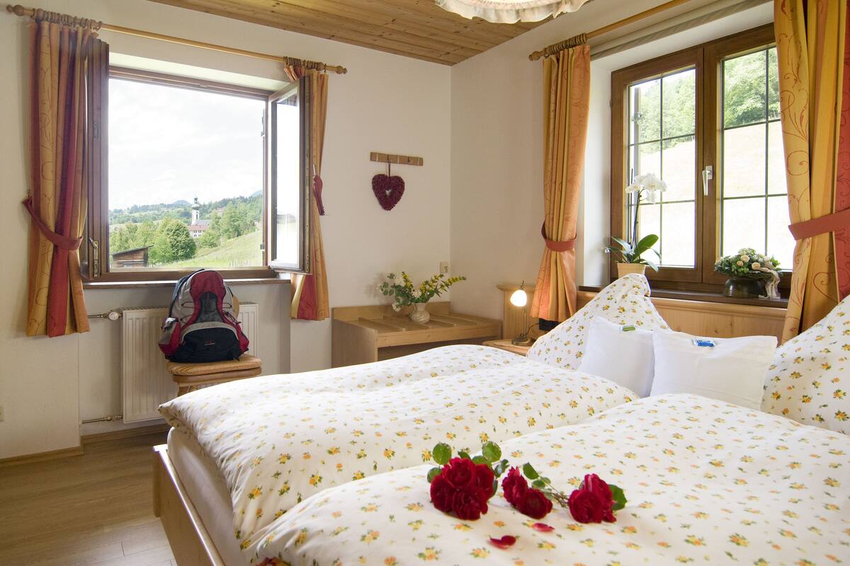 A bedroom with two beds, a window with curtains, a bedside table with a lamp, and a view of the countryside.