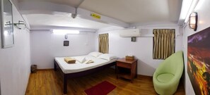 A compact hotel room with a single bed, a small bedside table, a green chair, an air conditioning unit, and a painting on the wall. - NZH Hostel (Yangon)