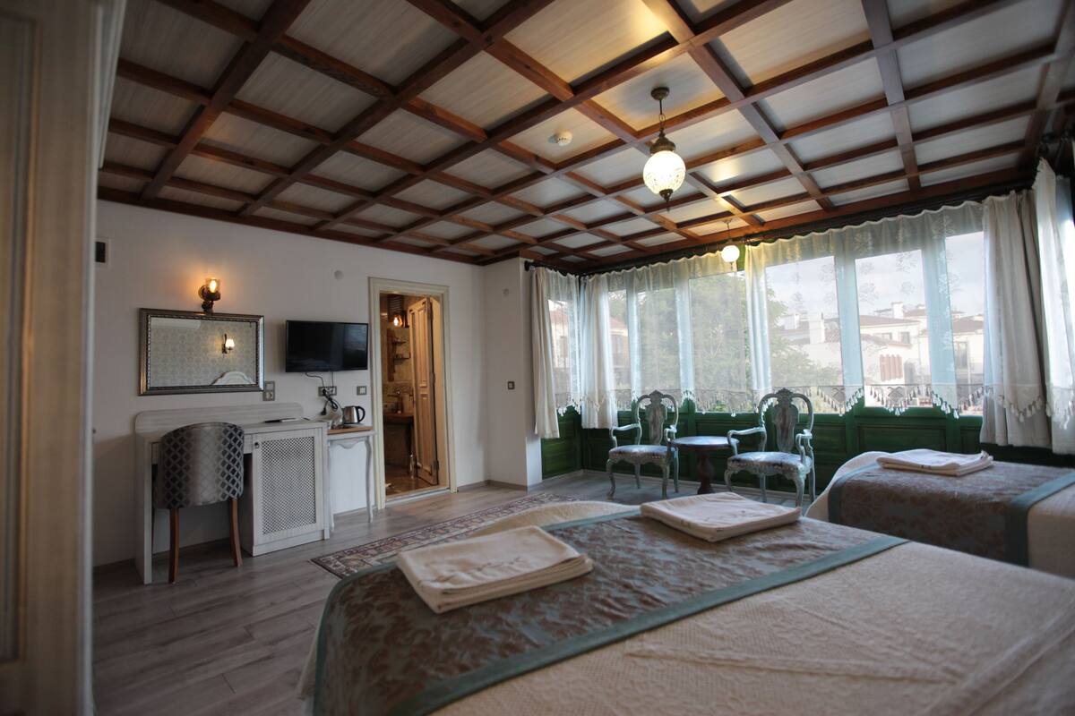 A hotel room with a wooden ceiling, a bed with a green bedspread, a desk with a chair, and a window with curtains.