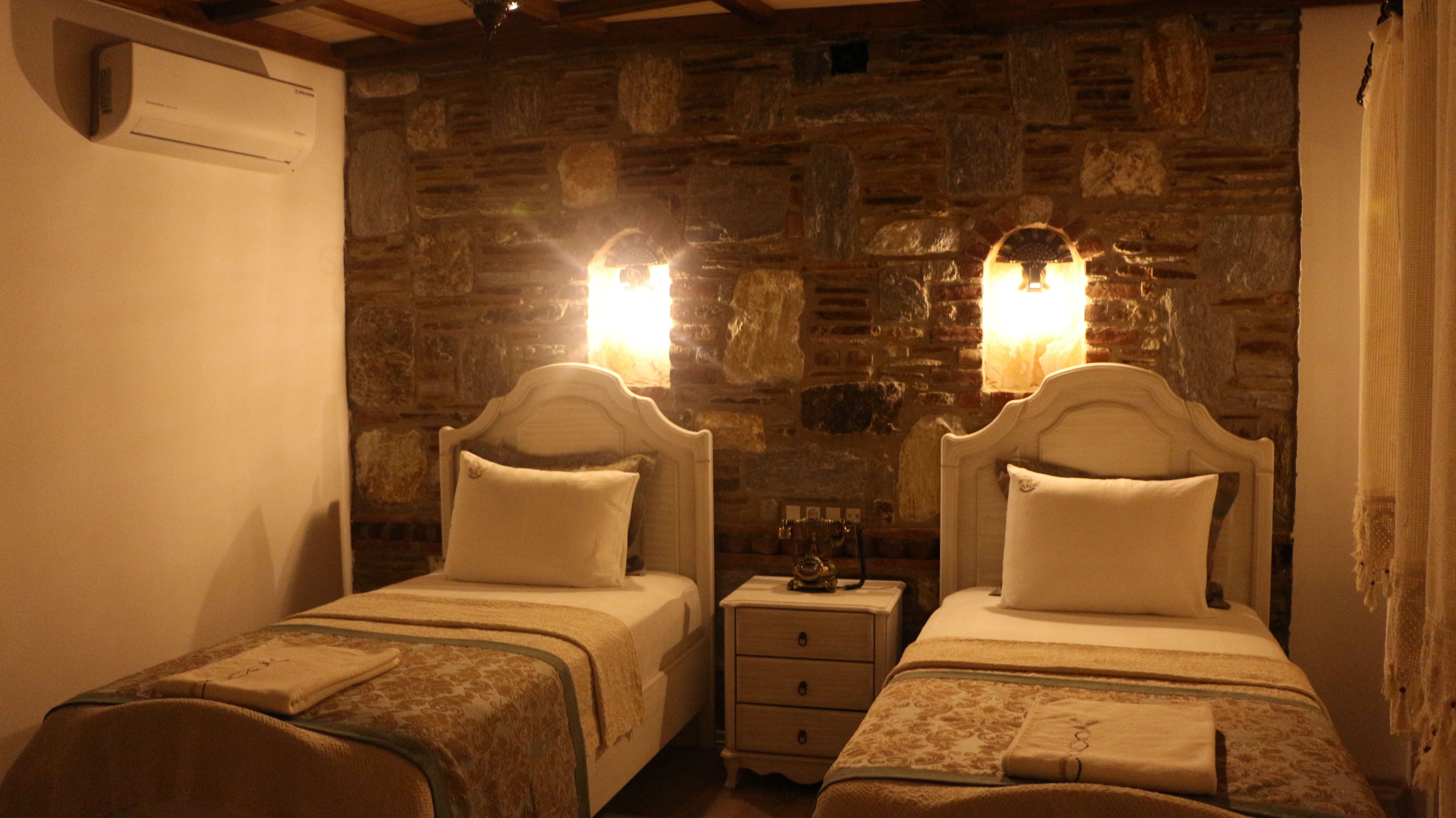 A room with two beds, each with a headboard and a bedside table. The walls are made of stone, and there is a small heater on the wall between the beds.