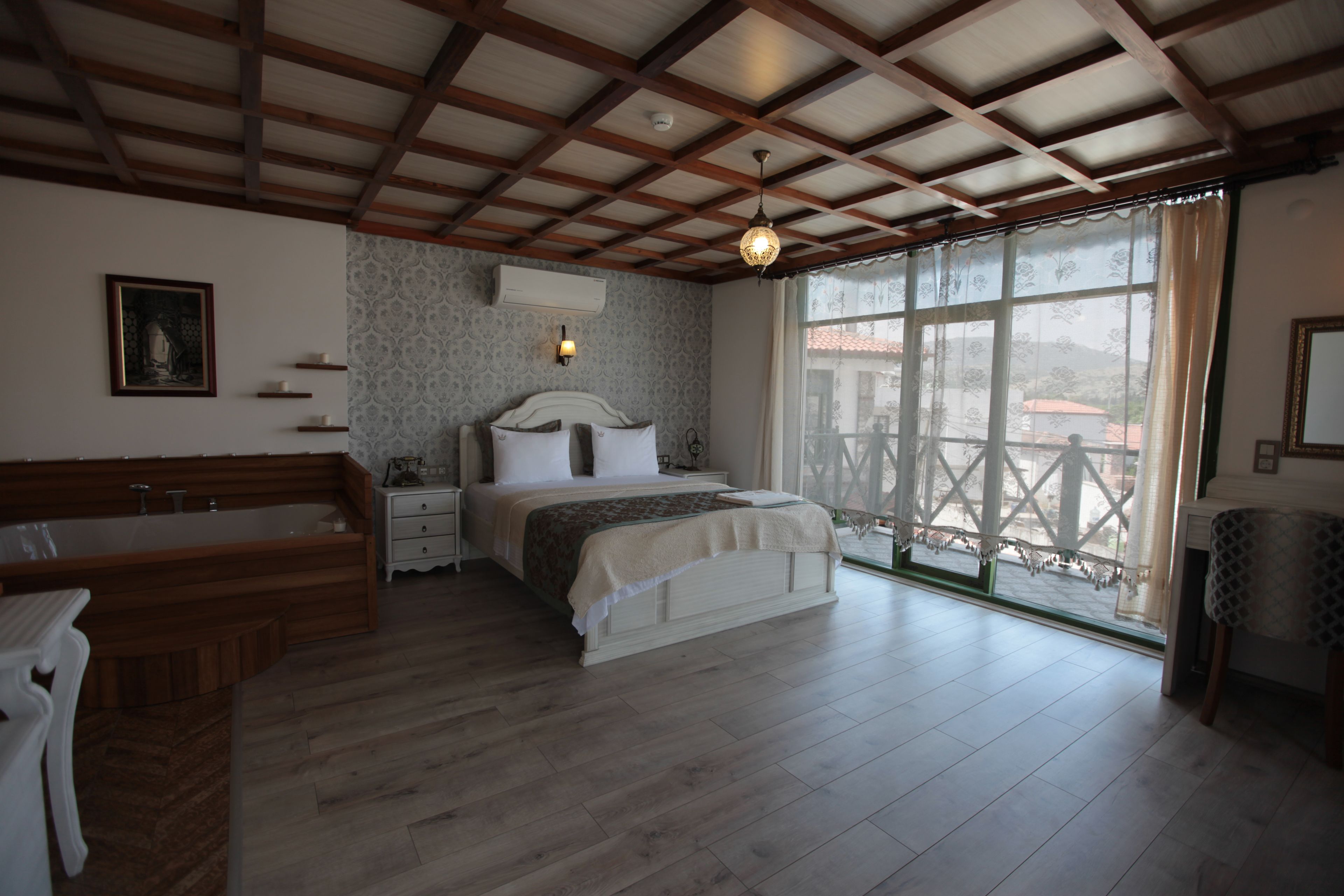 Un dormitorio con techo de madera, un ventanal con vistas, una cama con almohadas, una mesita de noche y una bañera de madera.