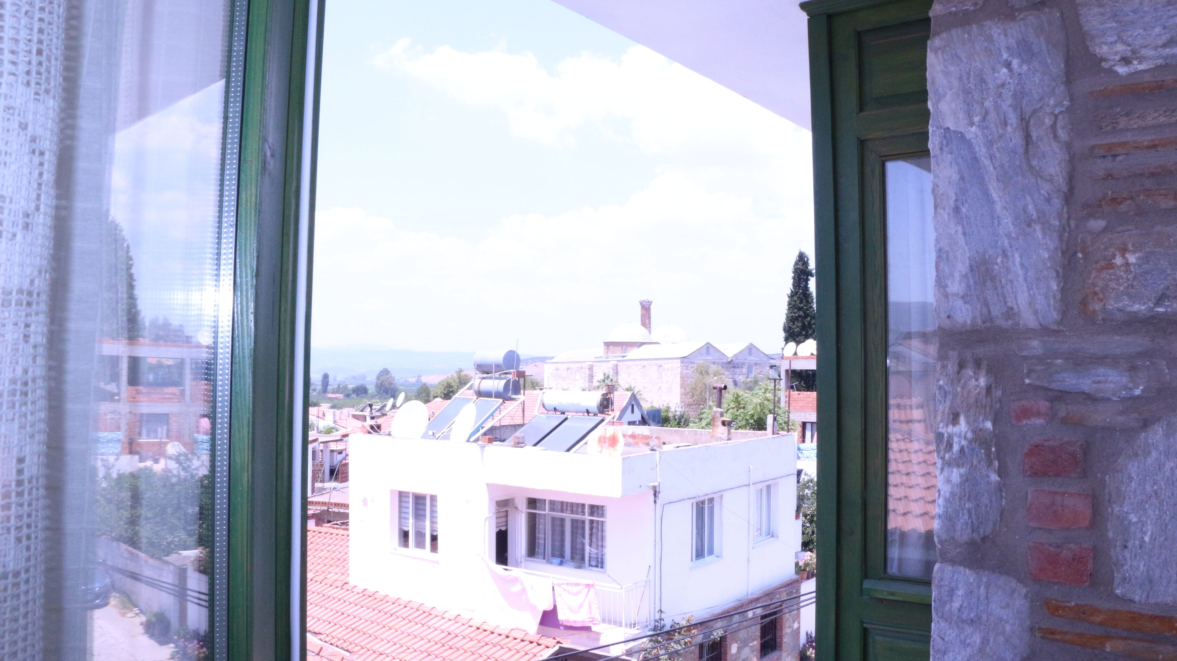Blick durch ein Fenster auf ein Gebäude mit rotem Ziegeldach, einem Balkon und einer Ziegelwand.