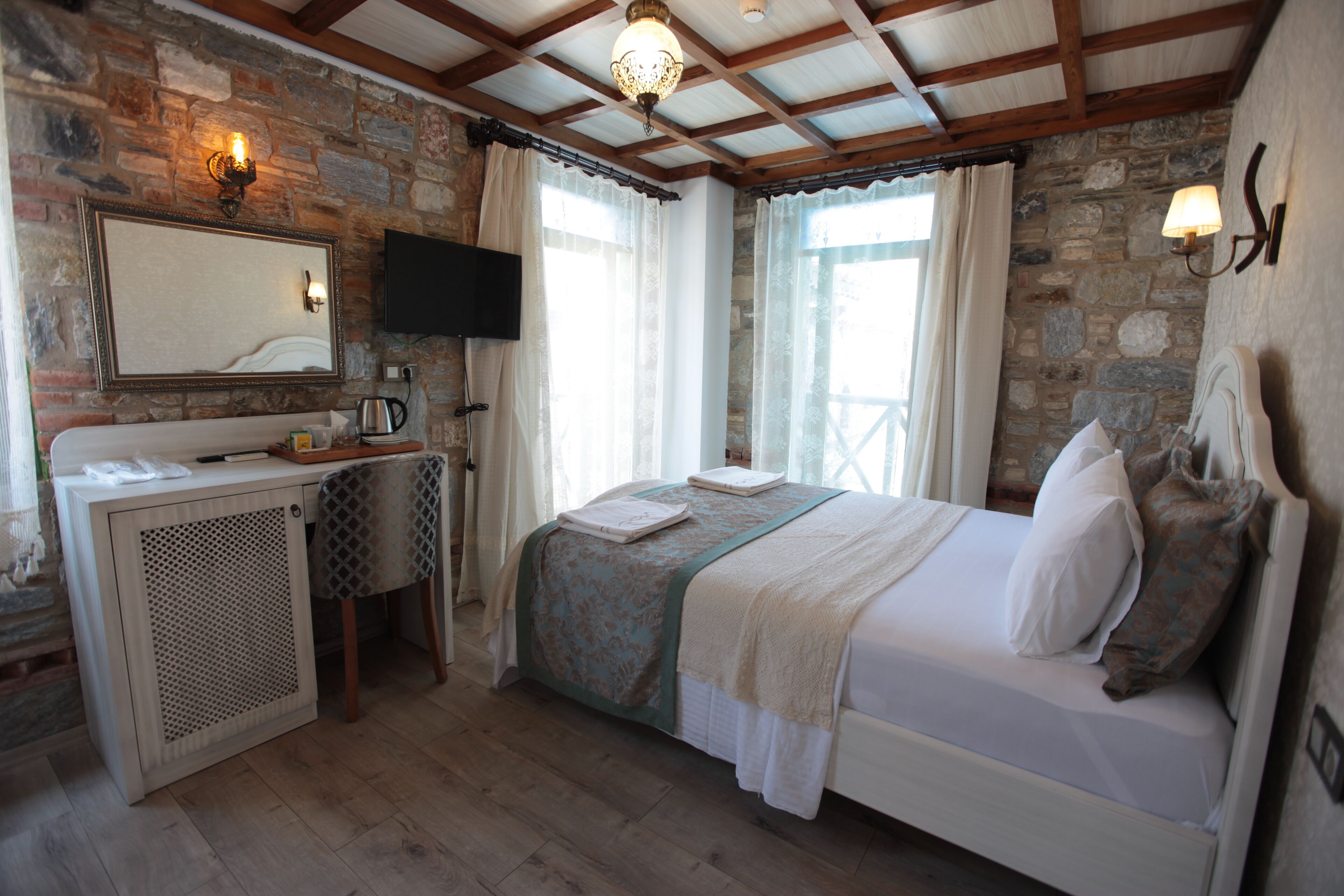 A bedroom with a stone wall, a bed with white linens, a bedside table with a lamp, and a television mounted on the wall.
