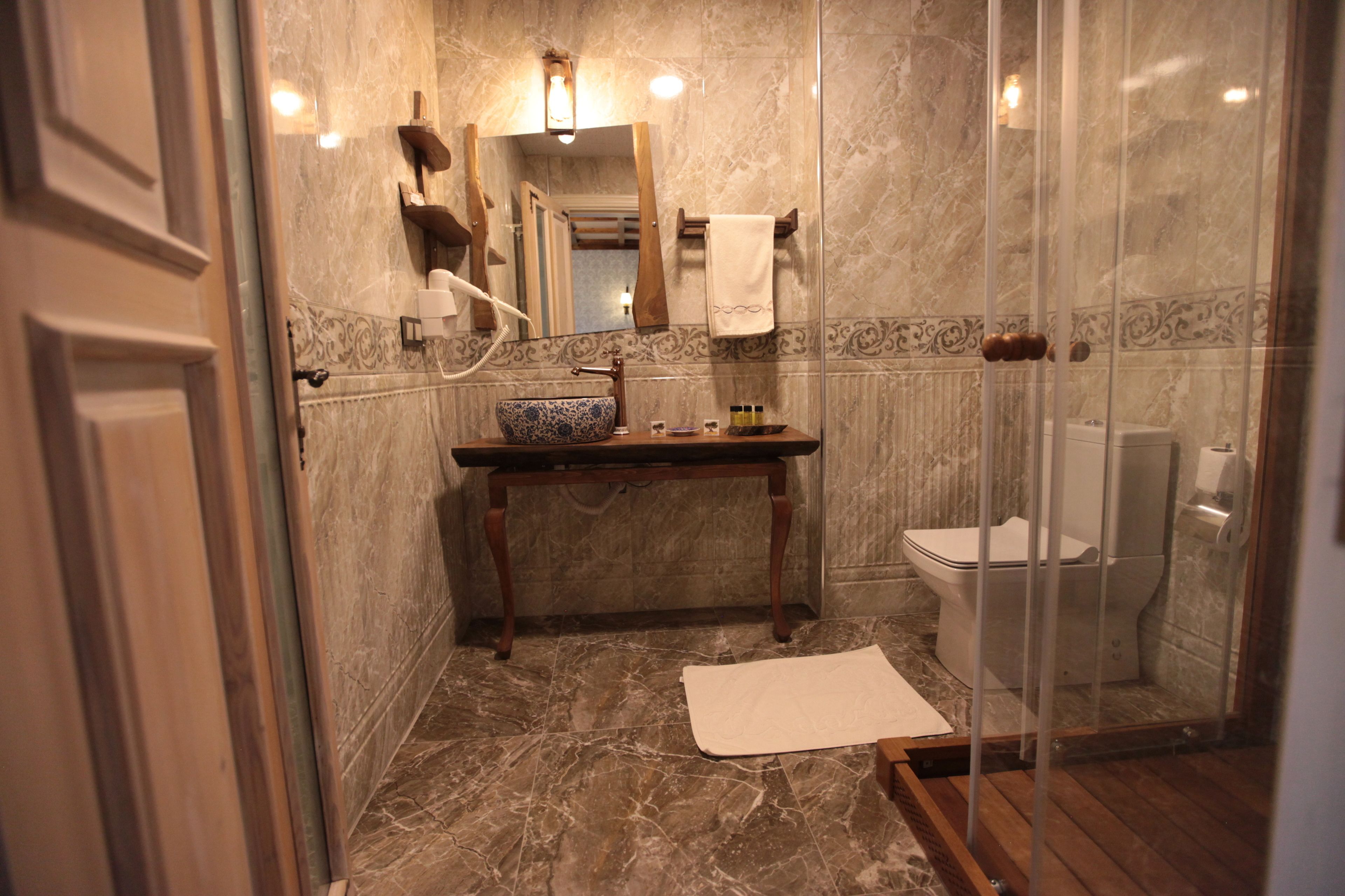 A bathroom with a marble floor, a wooden vanity, a sink with a blue bowl, a toilet, and a glass shower enclosure.