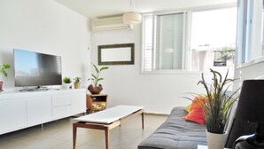 A modern living room with a flat-screen TV, a white cabinet, a wooden coffee table, a grey sofa with a cushion, and a potted plant. - Super Central Stunning Roof 2BR Apt (Tel Aviv)
