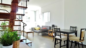 A modern living space with a staircase, a dining area with a table and chairs, a sofa with cushions, and a potted plant. - Super Central Stunning Roof 2BR Apt (Tel Aviv)