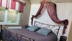 A bedroom with a bed, a nightstand, a window with a striped blind, and a decorative curtain. - Le Paddock du Val de Lys (Blessy)