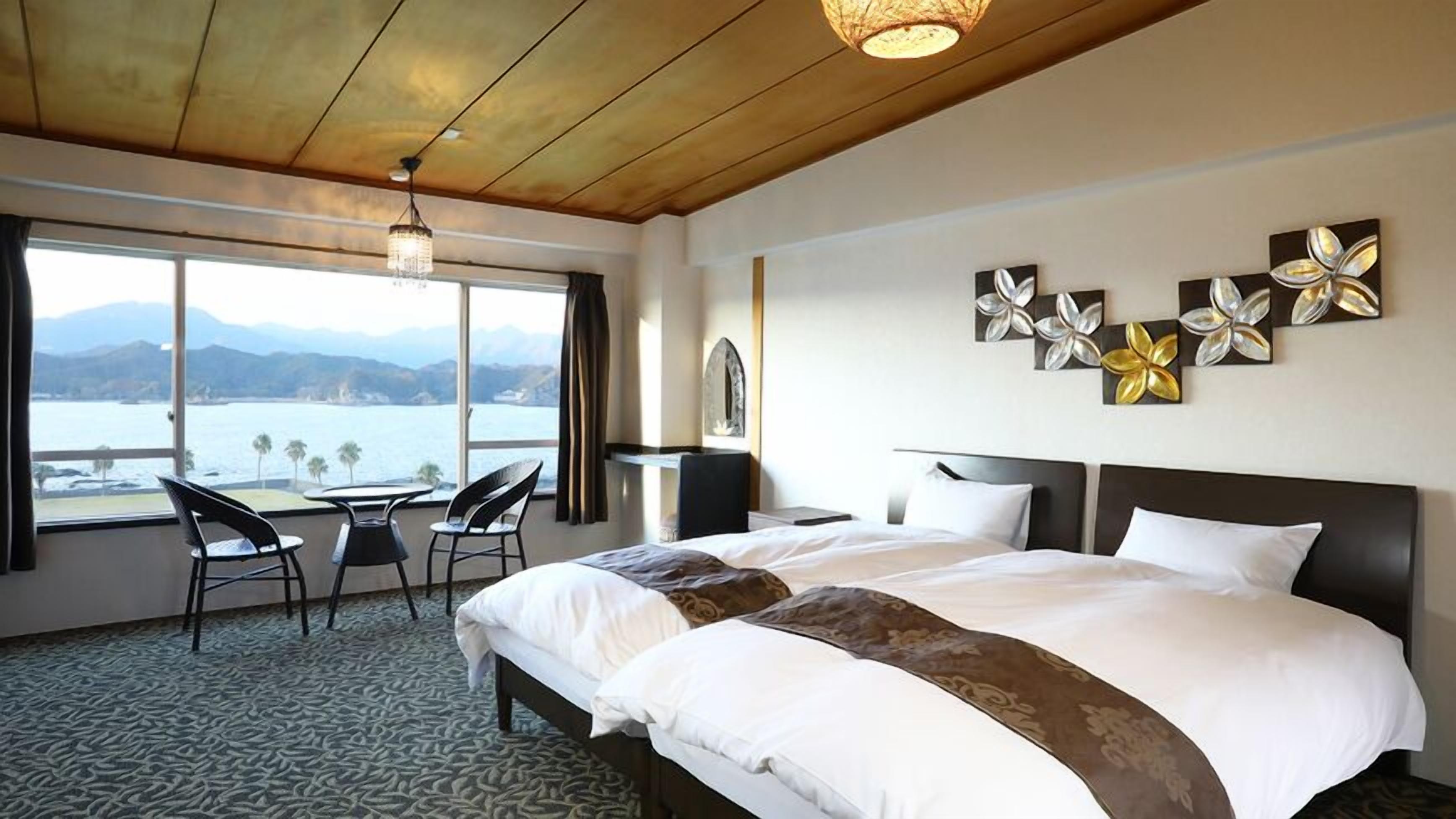 A hotel room with a bed, a desk, chairs, a view of the sea, and decorative wall art.