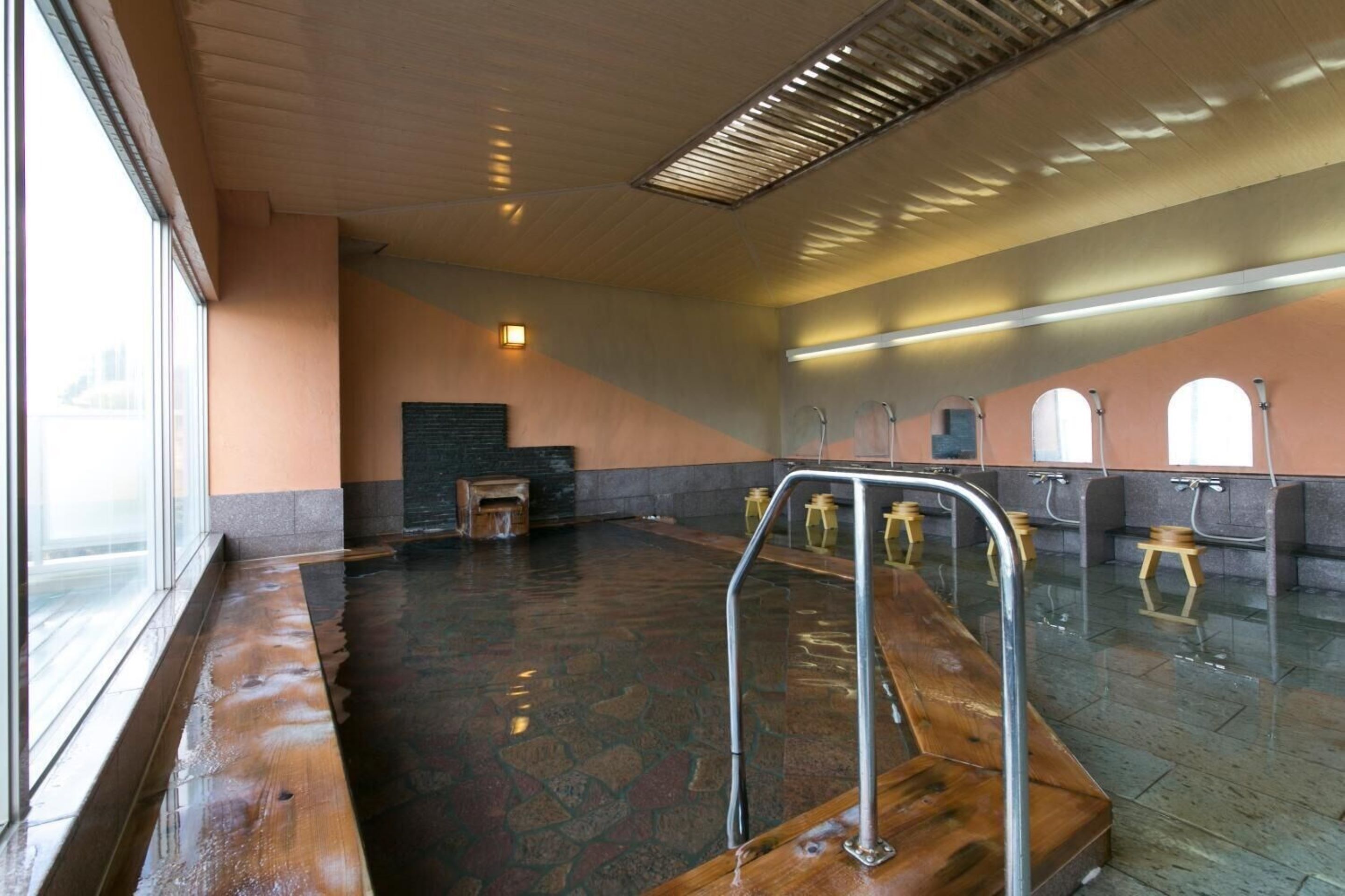 Baño público con piso mojado, un banco de madera y una barandilla.