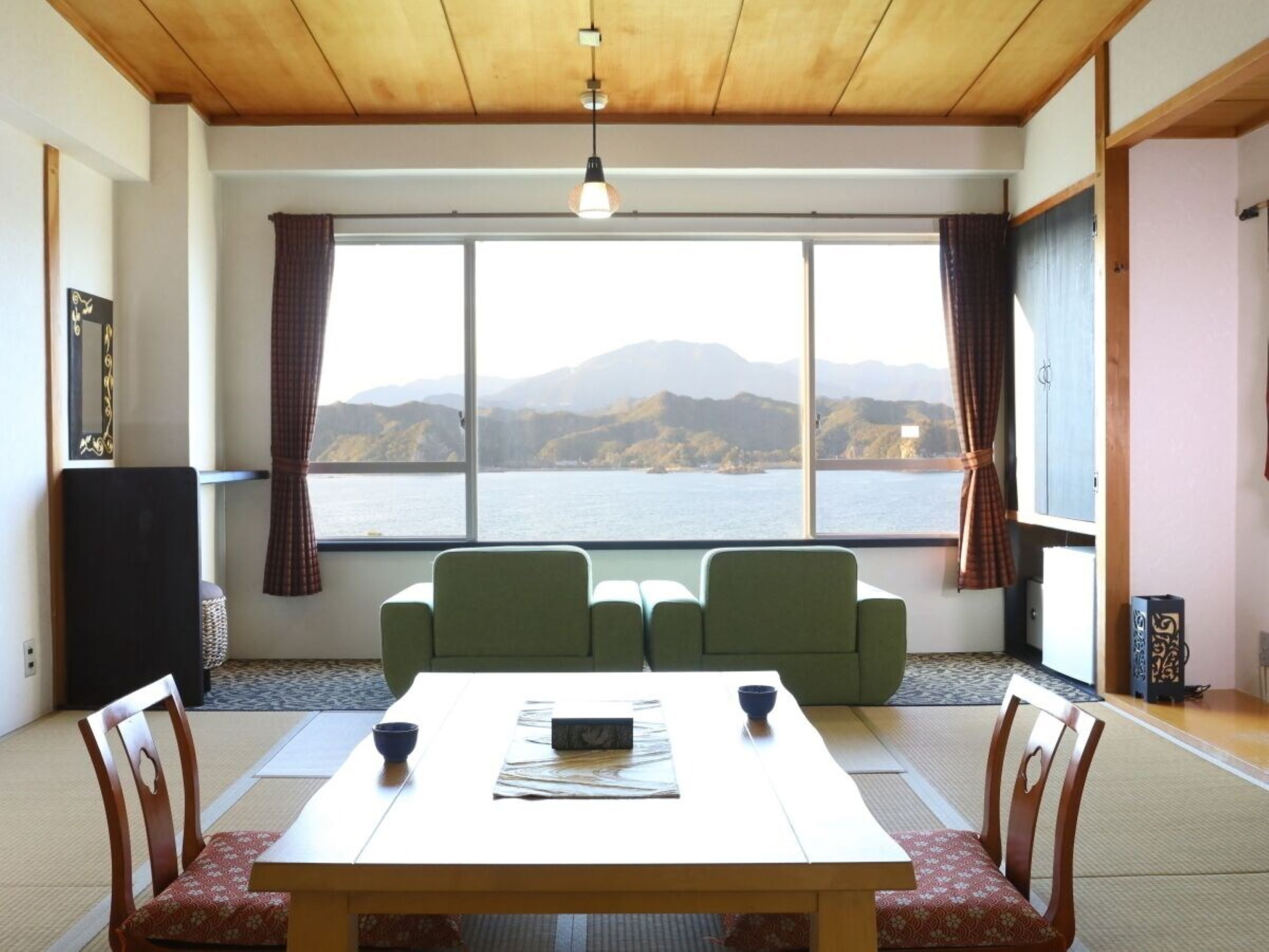 Une pièce avec un plafond en bois et une grande fenêtre donnant sur un paysage de montagnes et d’eau. On y trouve une table avec des chaises et un petit canapé.
