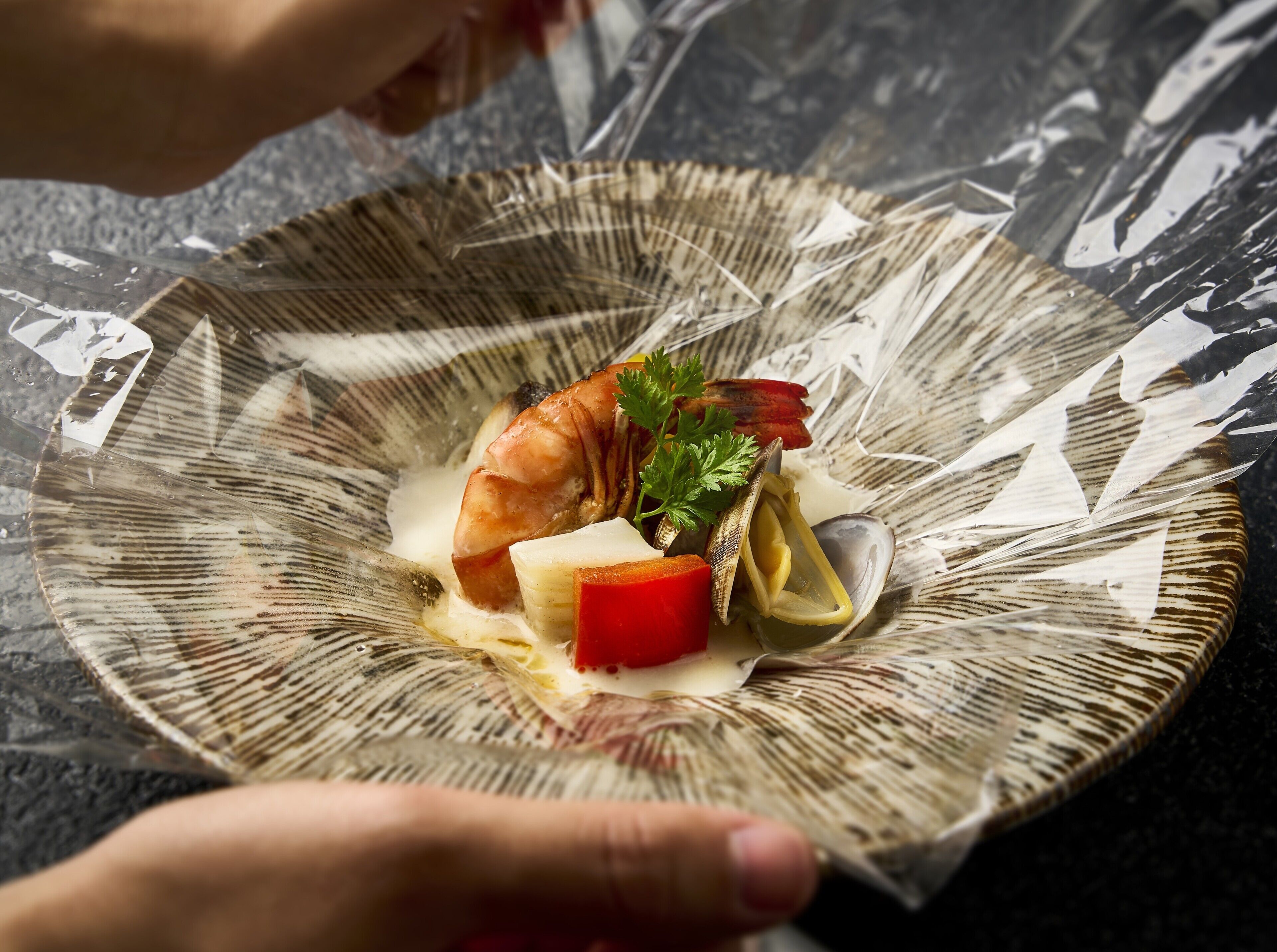 A plate with a shrimp, clams, and a red vegetable, possibly served on a textured ceramic dish.