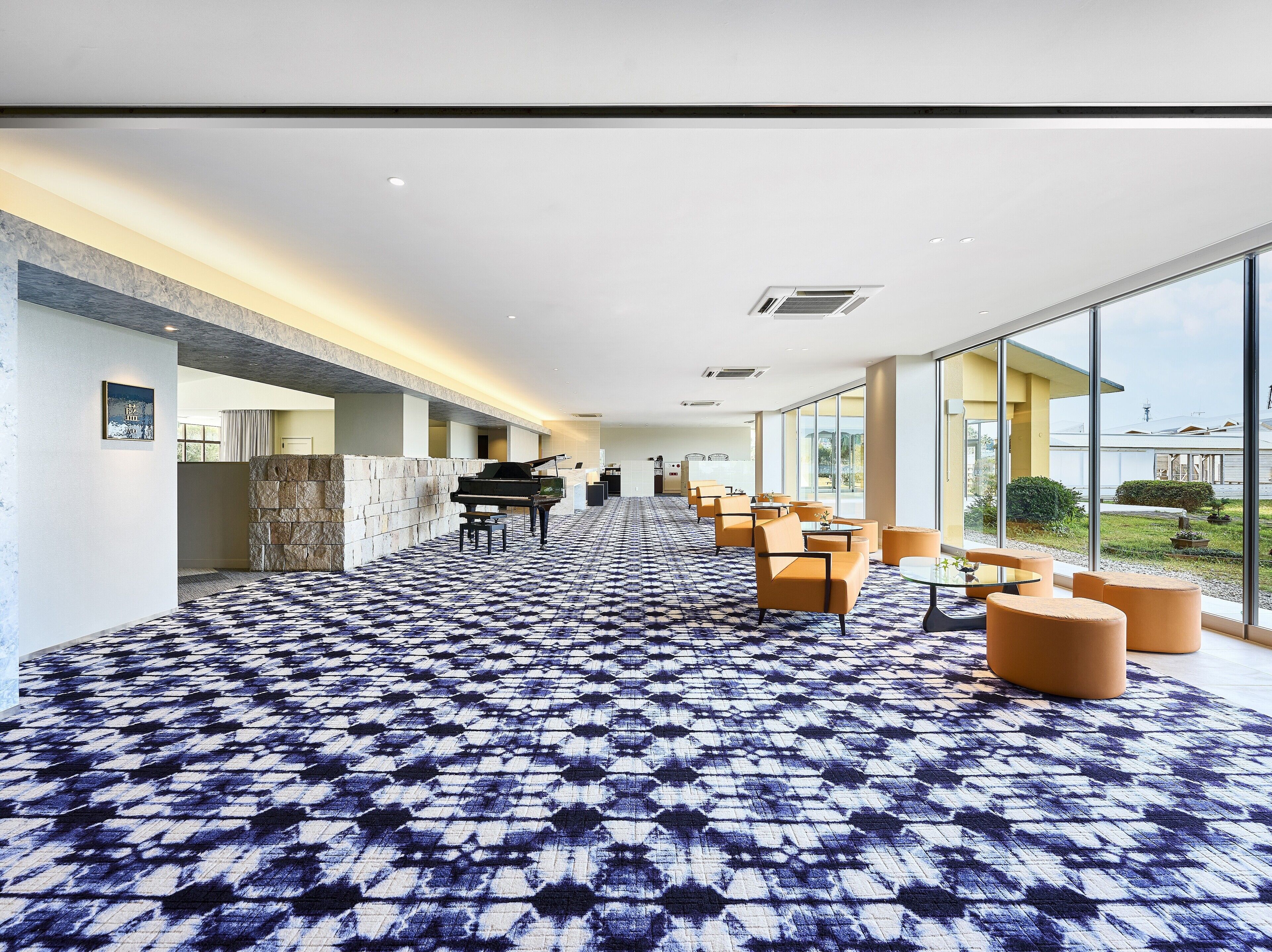 Un vestíbulo de hotel con alfombra estampada, un piano de cola y una zona de estar con amplias ventanas.