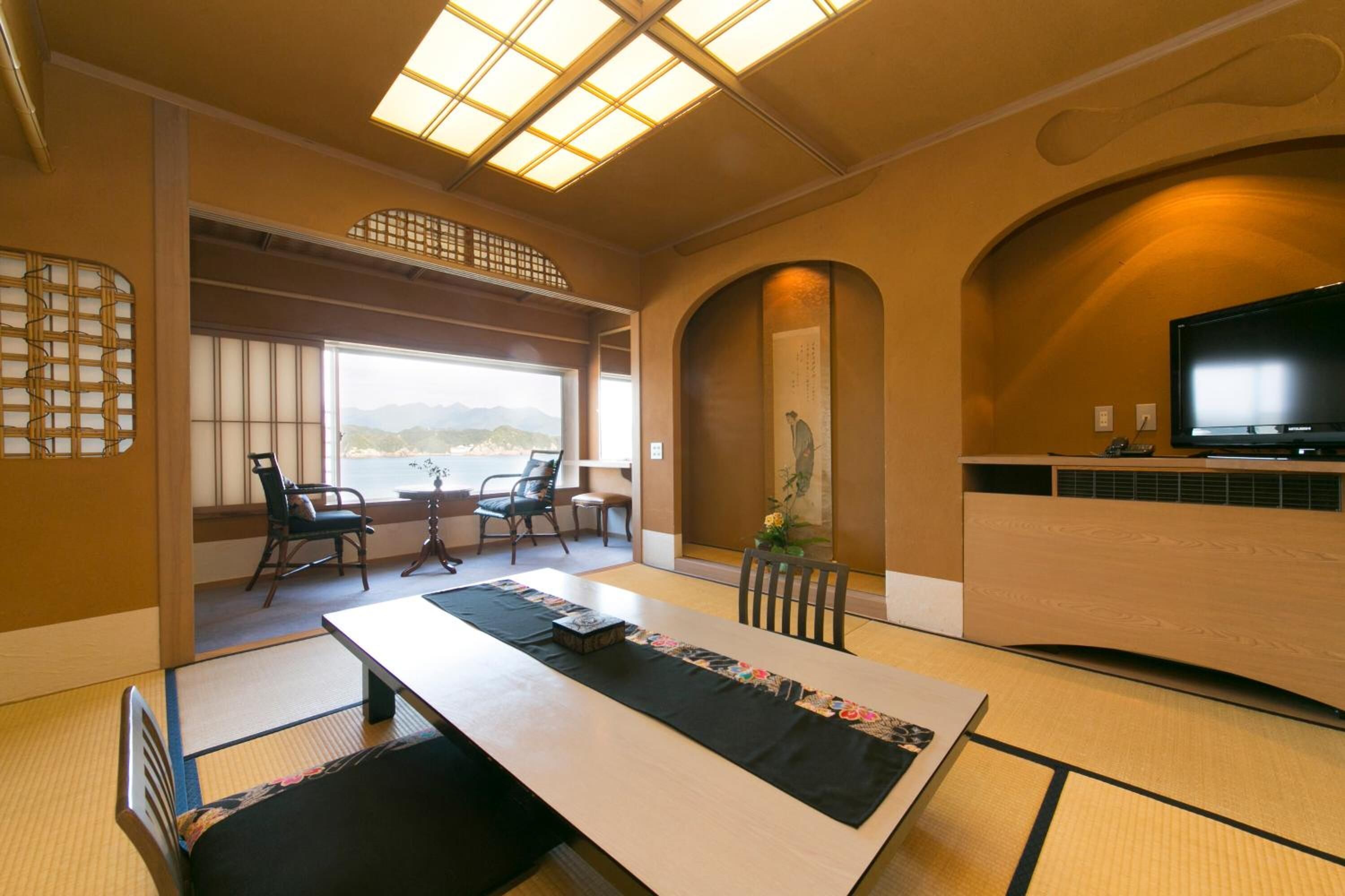 Um quarto japonês tradicional com tatames, uma mesa baixa com tapete preto, uma televisão e vista para montanhas através de grandes janelas.