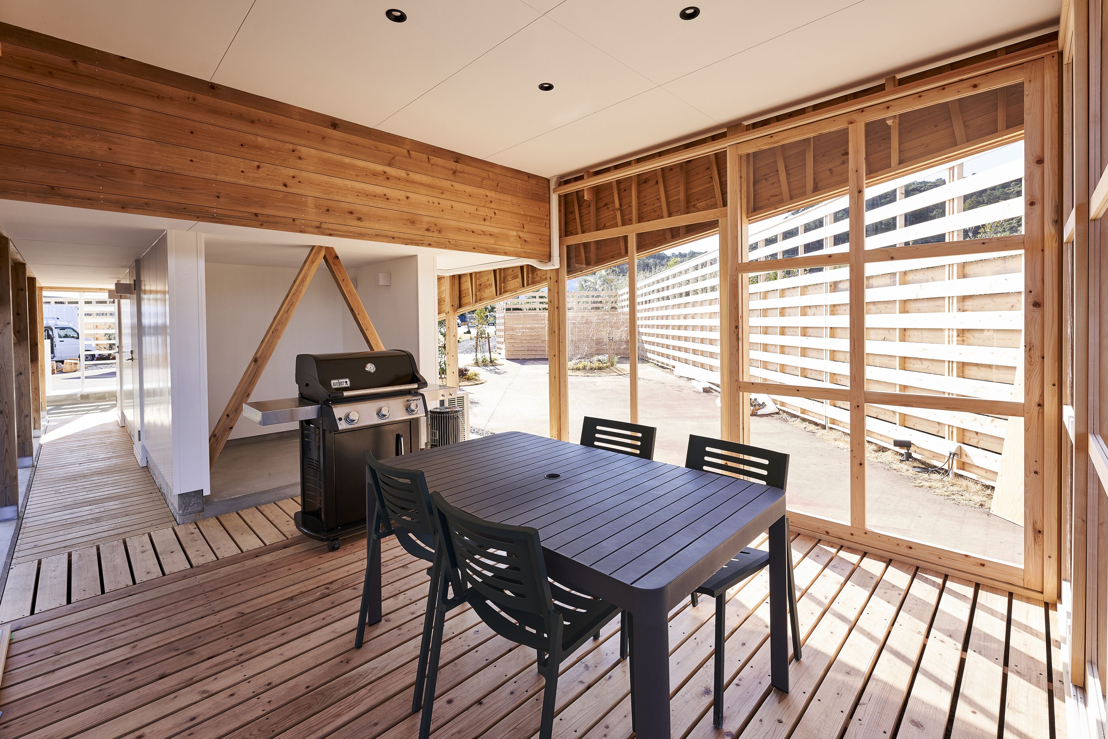 A wooden deck with a table and chairs, a grill, and a view of a fenced area.