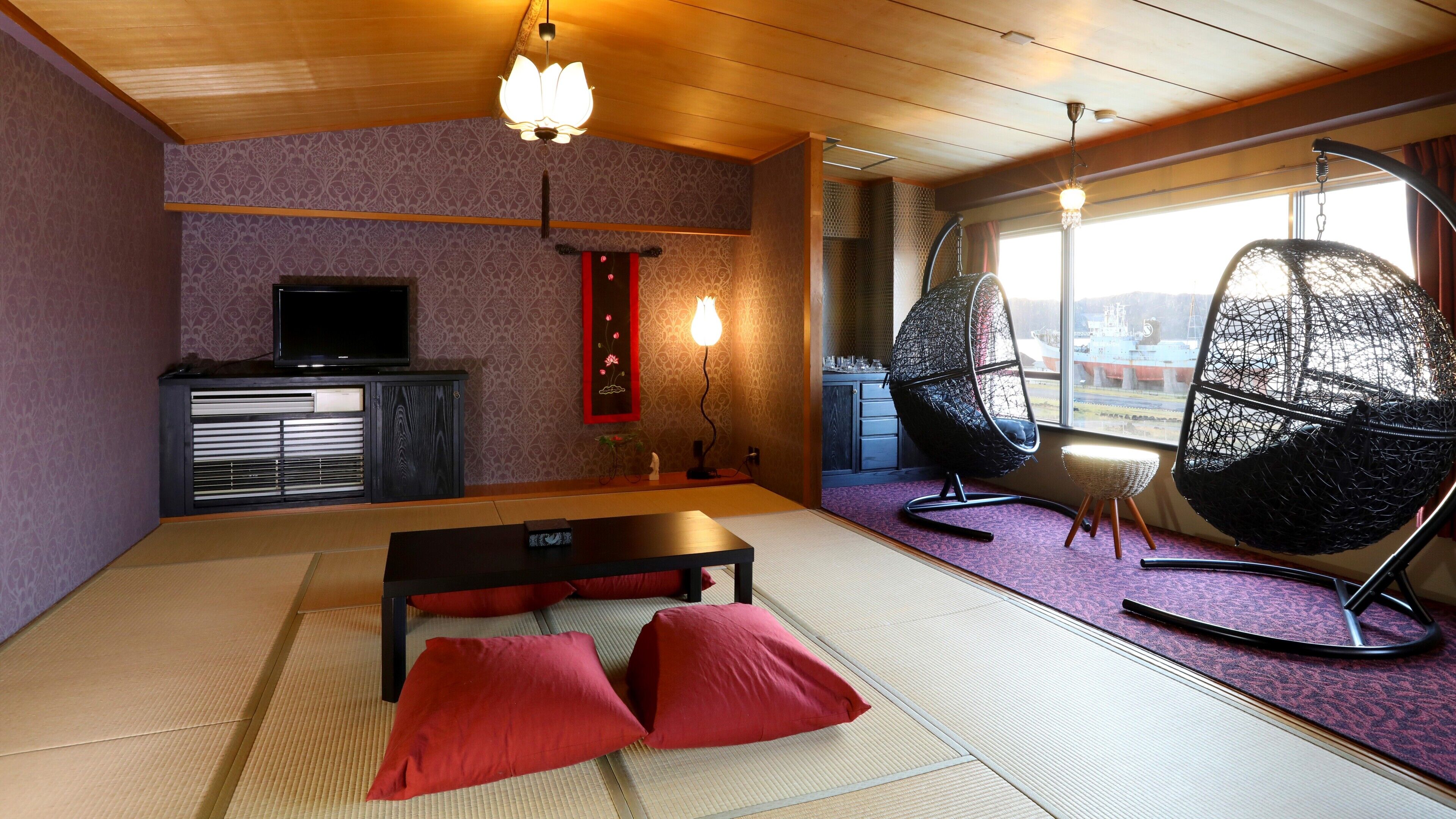 Una habitación con suelo de tatami, una mesa baja con cojines rojos, un televisor sobre un mueble y dos sillas colgantes.