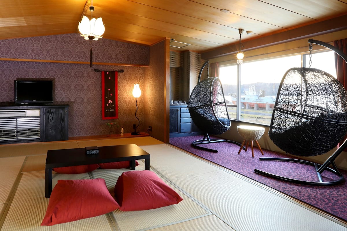 Una habitación con suelo de tatami, una mesa baja con cojines rojos, un televisor sobre un mueble y dos sillas colgantes.