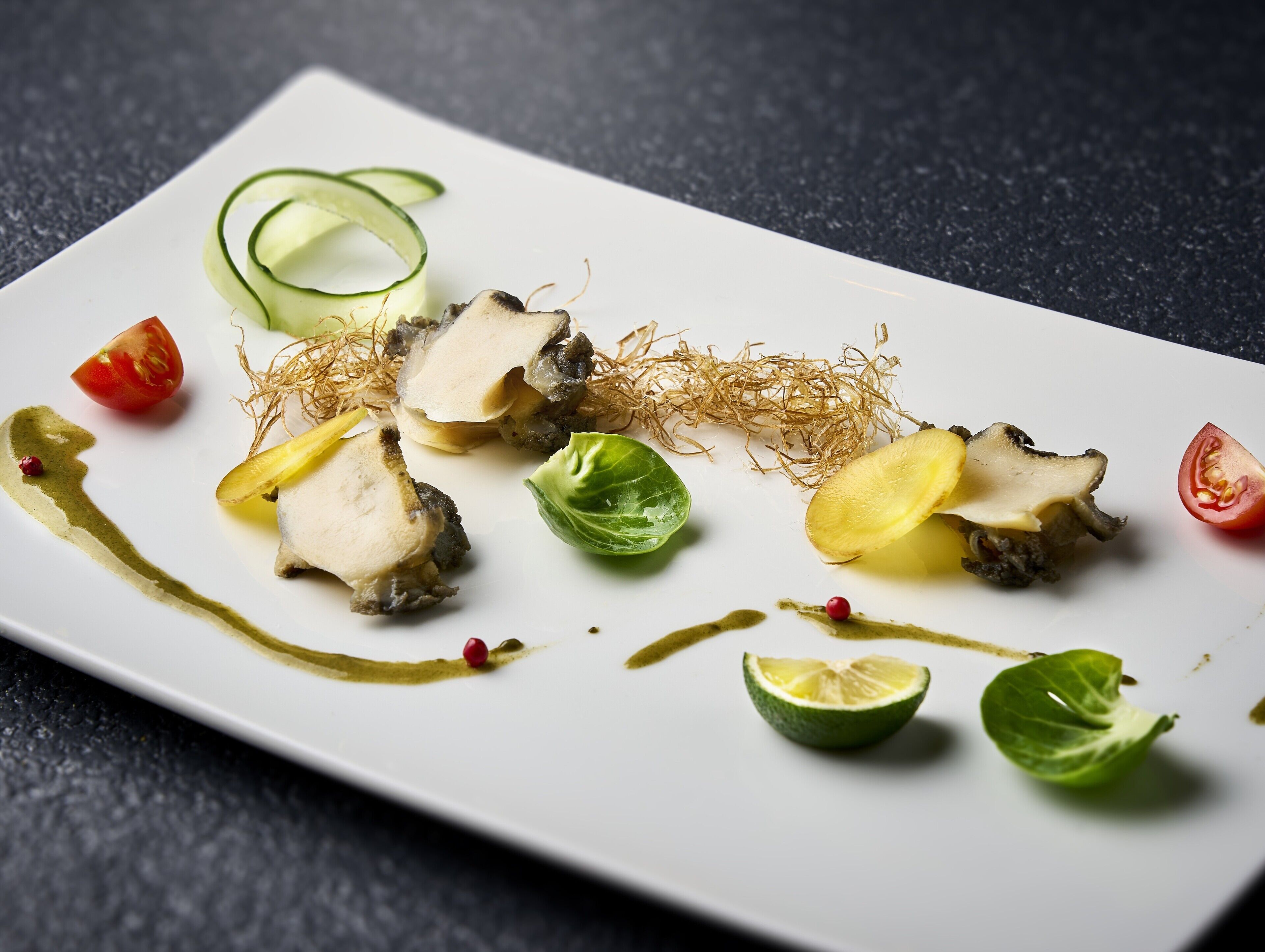 Un plato rectangular blanco con diversos alimentos, incluyendo rodajas de pepino, tomate y una verdura de hoja, sobre una superficie oscura y texturizada.