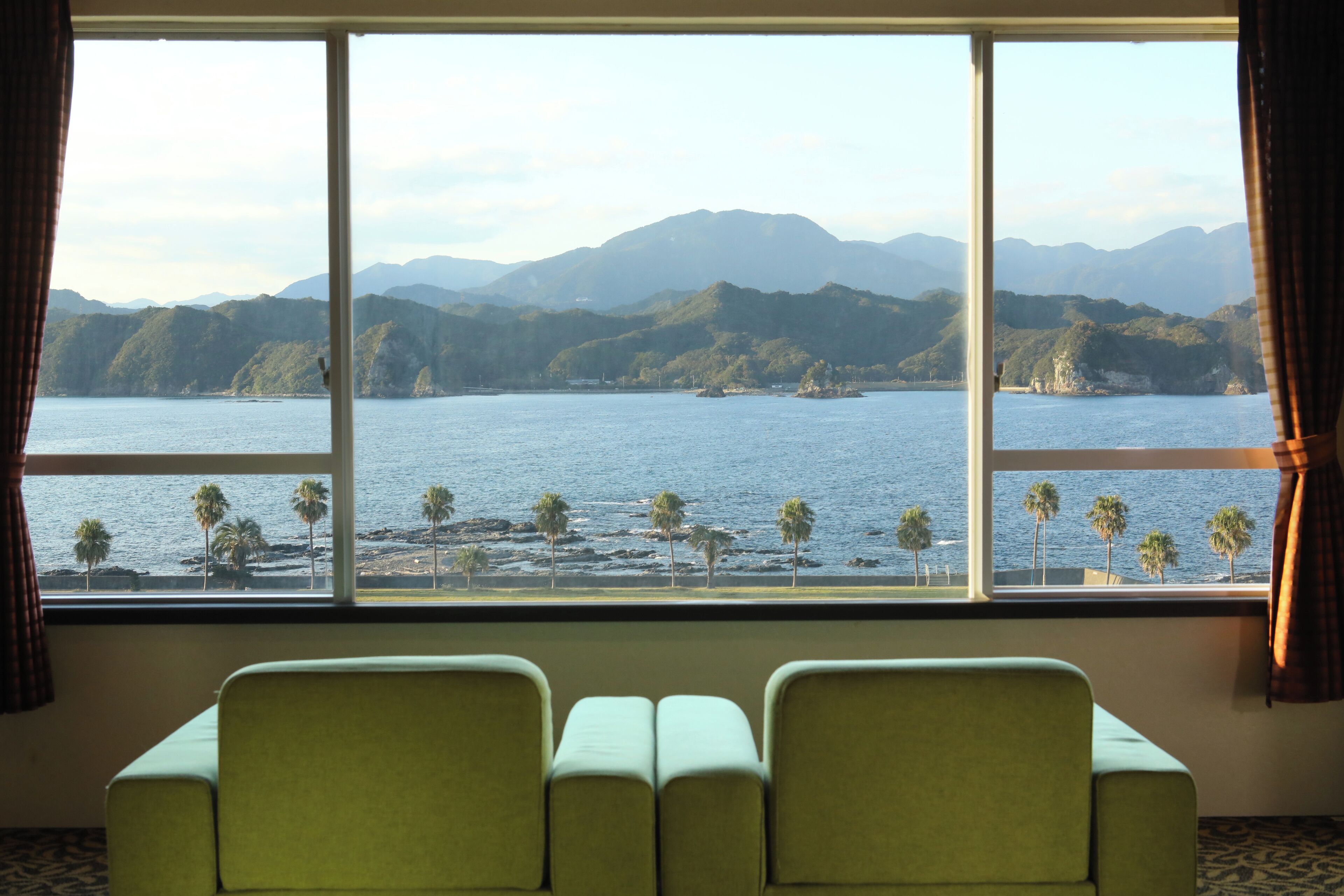 A room with a large window offering a view of a body of water and distant mountains. There are two green chairs in the foreground.