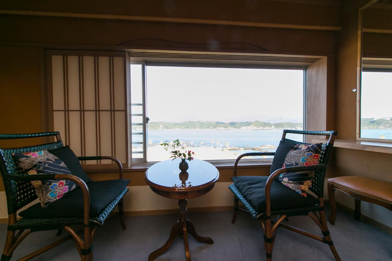 Een kamer met twee rieten stoelen, een klein rond tafeltje en uitzicht op zee door een groot raam.
