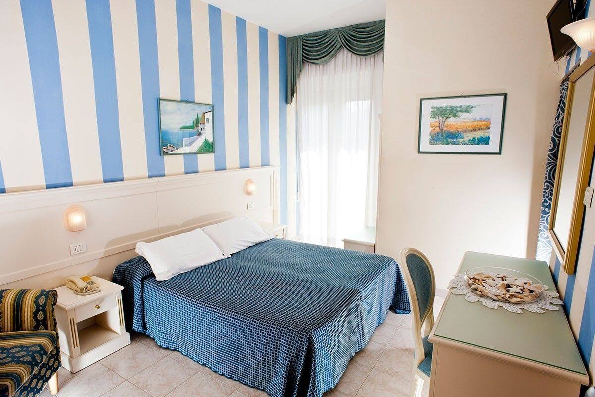 A hotel room with a bed, bedside table, chair, desk, and artwork on the walls.